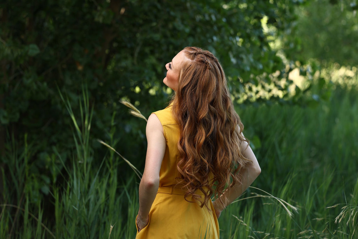 A woman with long, wavy reddish-brown hair and a braided crown stands in a lush green field wearing a yellow sleeveless dress.
